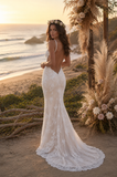 Woman in a white lace wedding dress standing on a beach with ocean and flowers in the background