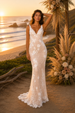 Woman in a white lace dress standing on a beach with sunset and floral decorations.