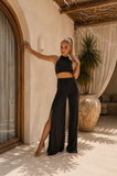 Woman in black outfit standing in a sunlit room with plants and furniture.