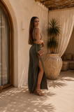 Woman in a green dress standing in a sunlit room with plants and furniture.