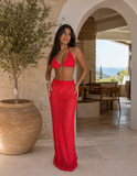 Woman in a red outfit Love Island USA standing in a sunlit outdoor setting with a view of the sea.