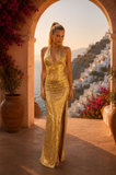 Woman in a gold dress standing in front of a scenic view with white buildings and flowers.